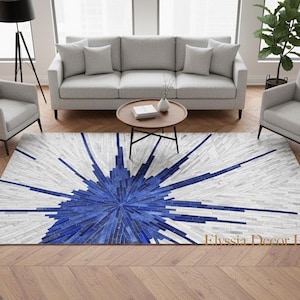 May include: A rectangular rug with a striking blue and white starburst design. The rug is placed on a hardwood floor and is surrounded by light gray furniture. The rug's design is centered with a burst of blue, radiating outwards.