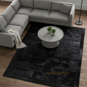 Pode incluir: Um tapete preto com um padrão geométrico está colocado sobre um chão de madeira. Um sofá cinza em forma de L e uma mesa de centro redonda cinza claro com duas pequenas plantas em vasos estão no fundo. O texto "Elyssia Decor Home" é visível no tapete.