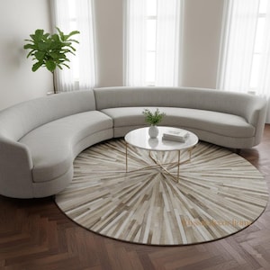 May include: A curved, light gray sectional sofa with a round marble-topped coffee table and a patterned rug. The rug features a radial design in shades of beige and brown. A potted plant is visible in the corner, and the room has large windows.