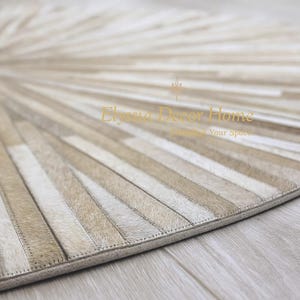 May include: A beige and cream-colored round rug with a radial pattern. The rug is made of rectangular pieces of material sewn together, creating a sunburst design. The rug has a stitched border and is placed on a light-colored wooden floor.