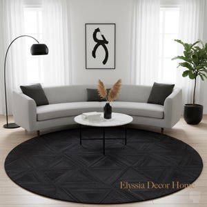 May include: A modern living room featuring a light gray curved sofa with dark gray throw pillows. A round marble coffee table sits on a geometric patterned black rug. A black floor lamp and a potted plant add to the decor.
