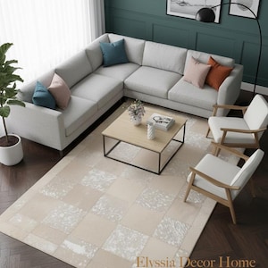 May include: A contemporary living room arrangement with a light grey corner sofa and colourful cushions. A square wooden coffee table rests on a beige and white patterned rug. Two white armchairs and framed art complete the scene.