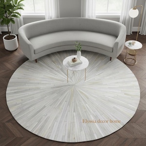 Può includere: Un moderno salotto con un divano curvo grigio, due tavolini con piano in marmo e un grande tappeto rotondo con un motivo a raggiera. Il tappeto è principalmente bianco e grigio. La stanza ha una palette di colori neutri ed è ben illuminata.
