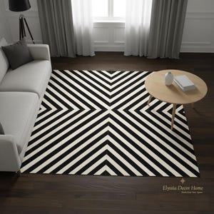 Könnte beinhalten: Ein schwarz-weiß gemusterter Teppich mit geometrischem Design. Das Muster besteht aus sich wiederholenden schwarzen und weißen Streifen, die sich in der Mitte treffen. Der rechteckige Teppich liegt auf einem dunklen Holzboden.