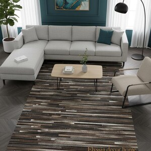 May include: A modern living room featuring a light gray sectional sofa, a wooden coffee table, and a patterned rug. The rug has a striped design in shades of brown, gray, and white. A painting with teal and gold accents hangs on the wall.
