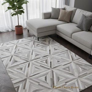 May include: Geometric patterned area rug in shades of gray and white. The rug features a repeating triangular design. The rug is placed on a dark wood floor in a living room setting. A gray sectional sofa and a potted plant are also visible.