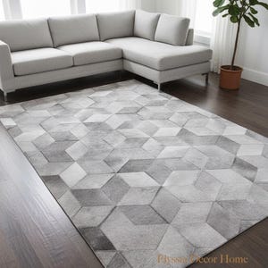 Könnte beinhalten: Ein grau gemusterter Teppich mit einem 3D-Würfeldesign. Der Teppich liegt auf einem dunklen Holzboden, im Hintergrund ein graues Ecksofa. Rechts ist eine Topfpflanze zu sehen. Das Design des Teppichs besteht aus verschiedenen Grautönen.