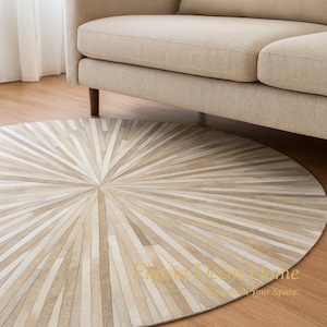 May include: A round, beige and cream-colored rug with a radial pattern, placed on a wooden floor. The rug is positioned beneath a beige sofa with wooden legs, creating a warm and inviting living space.