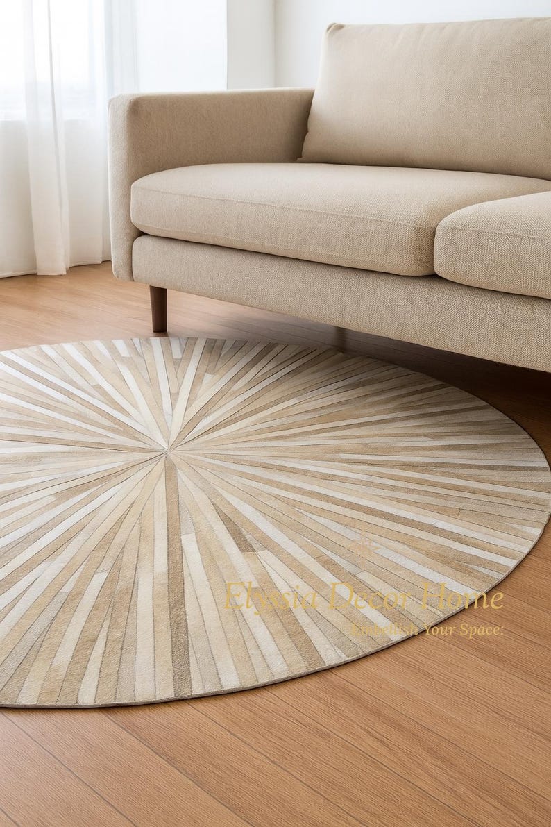 May include: A round, beige and cream-colored rug with a radial pattern, placed on a wooden floor. The rug is positioned beneath a beige sofa with wooden legs, creating a warm and inviting living space.