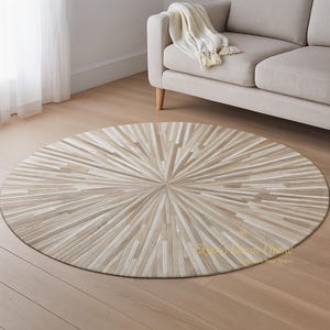 May include: A round rug with a sunburst design. The rug is made of light beige and cream-colored rectangular pieces arranged in a radial pattern. The rug is placed on a light wood floor, with a beige sofa in the background.