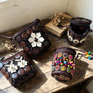 May include: Four handcrafted bags on a wooden surface. Two square bags feature dark brown discs and white flower accents. A cylindrical bag is adorned with colourful beads and wooden flower shapes. Another cylindrical bag has a lid and is decorated with brown and white discs.