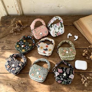 Può includere: Sette piccole custodie in tessuto trapuntato per auricolari wireless, ciascuna dotata di manico superiore, chiusura con bottone a pressione e vari motivi floreali o botanici in rosa, verde, bianco, blu navy e nero, con auricolari bianchi.