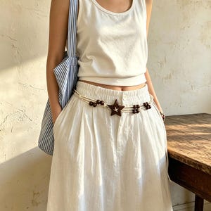 May include: A white tank top and white skirt paired with a blue and white striped tote bag. A waist belt features a dark brown star-shaped charm and dark brown wooden beads strung on thin, light-colored cords that wrap around the waist.