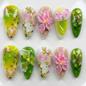 May include: A set of ten artificial nails with floral designs. The nails feature a variety of colors, including green, pink, and white, with 3D flower embellishments and water droplet accents. The nails are arranged on a white surface.