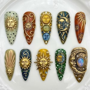 May include: A collection of ten intricately designed artificial nails. Each nail features a unique celestial design, including sun and moon faces, in gold, bronze, and other colors. Some nails have decorative swirls and dots.