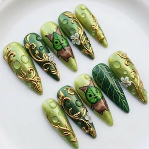 Puede incluir: Un conjunto de diez u&ntilde;as artificiales largas en forma de almendra. Las u&ntilde;as presentan gradientes de tonos verdes, con dise&ntilde;os de filigrana dorada, hojas y flores. Algunas u&ntilde;as tienen un personaje de hoja de dibujos animados. Las u&ntilde;as est&aacute;n dispuestas sobre un plato blanco.