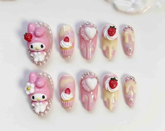 May include: A set of ten artificial nails with a pink and white theme. The nails feature various designs, including a cartoon character, cupcakes, hearts, and strawberries. Some nails are adorned with rhinestones and pearl accents.