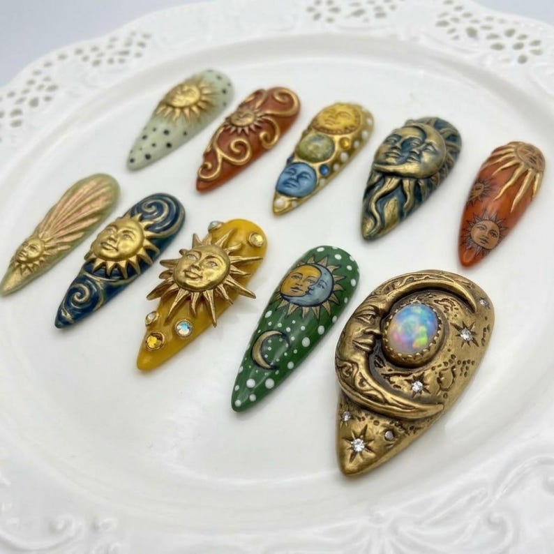 May include: A collection of ten almond-shaped nail art designs. Each nail features celestial-themed art, including suns, moons, and stars, in gold, blue, green, and orange hues. Some nails have textured details and embellishments.