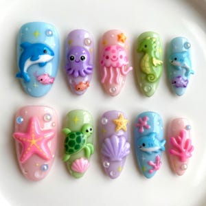 May include: A set of ten artificial nails with a sea life theme. Each nail features a different 3D design, including a dolphin, octopus, jellyfish, seahorse, starfish, turtle, seashell, and coral, in pastel colors. The nails are oval-shaped.