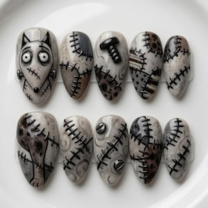 May include: A set of ten press-on nails featuring a grey and white color palette with a Frankenstein-inspired theme. The designs include stitched patterns, a cartoon creature face, metal bolt accents, and various textured patches in shades of grey.