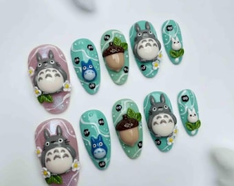 May include: Set of custom-designed nail art featuring characters from the animated film, "My Neighbor Totoro." The nails are painted in various colors, including pink and teal, and adorned with 3D Totoro figures, acorns, and other decorative elements.