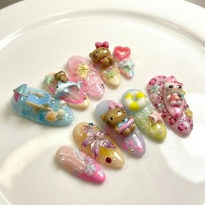 Puede incluir: Una colecci&oacute;n de u&ntilde;as artificiales decoradas. Los dise&ntilde;os incluyen osos de peluche, corazones, un delf&iacute;n, una silla de playa y un salvavidas. Las u&ntilde;as est&aacute;n pintadas en varios colores, incluyendo rosa, azul, amarillo y blanco, con purpurina y adornos.