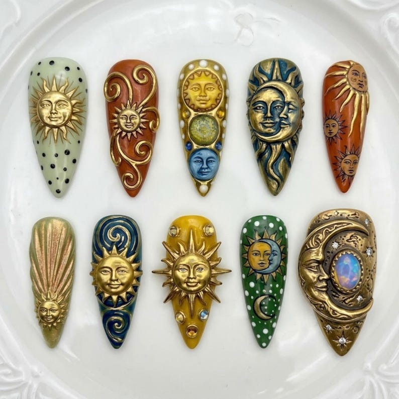 May include: A collection of ten intricately designed artificial nails. Each nail features a unique celestial design, including sun and moon faces, in gold, bronze, and other colors. Some nails have decorative swirls and dots.