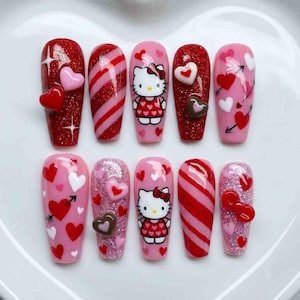 May include: A set of ten artificial nails with Valentine's Day themed designs. The nails feature red glitter, pink hearts, and a Hello Kitty design. Some nails have stripes, arrows, and 3D heart accents.