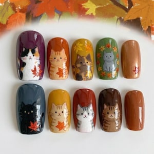 May include: A set of ten press-on nails featuring various cat illustrations and autumn leaf motifs. The designs include a calico, orange tabby, tortoiseshell, gray cat, black cat, and brown tabby, painted on backgrounds of purple, orange, yellow, green, blue, and brown.