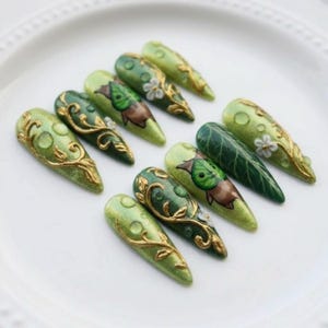 Puede incluir: Diez u&ntilde;as postizas en forma de almendra, en tonos verdes variados. Las u&ntilde;as est&aacute;n decoradas con filigranas doradas, dise&ntilde;os de hojas y peque&ntilde;as flores blancas. El dise&ntilde;o est&aacute; inspirado en la naturaleza, con un acabado elegante y detallado.