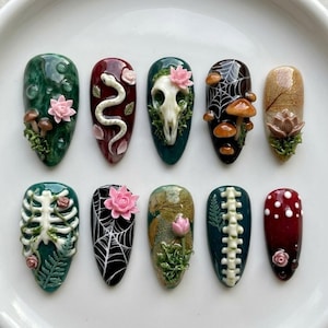 May include: A set of ten press-on nails featuring intricate 3D designs including a white snake, a skull, mushrooms, spiderwebs, a skeleton ribcage, a spine, a fern, and pink lotus flowers on dark green, red, black, and tan backgrounds.
