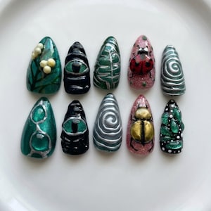May include: A set of ten almond-shaped press-on nails featuring diverse designs including a green leaf pattern with white berries, a black eye motif, silver swirls, a red ladybug on pink glitter, a gold beetle, and green textured accents.