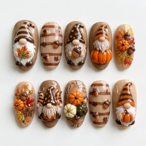 May include: A set of ten press-on nails featuring autumn-themed designs. The collection includes 3D gnomes with striped or polka-dot hats, pumpkins, acorns, and colorful fall leaves in shades of orange, brown, cream, and green on a tan base.