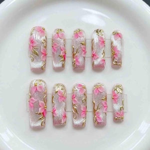 May include: Set of ten artificial nails with a glittery silver base, adorned with pink flower accents and gold filigree. The nails are rectangular shaped and arranged on a white surface.