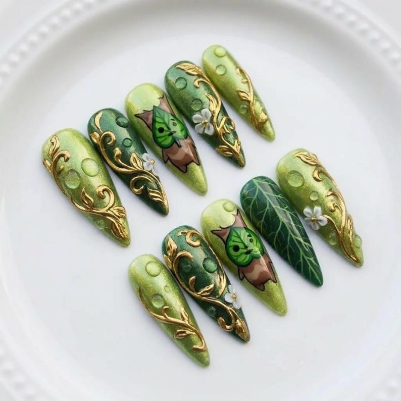 Puede incluir: Un conjunto de diez u&ntilde;as artificiales largas en forma de almendra. Las u&ntilde;as presentan gradientes de tonos verdes, con dise&ntilde;os de filigrana dorada, hojas y flores. Algunas u&ntilde;as tienen un personaje de hoja de dibujos animados. Las u&ntilde;as est&aacute;n dispuestas sobre un plato blanco.