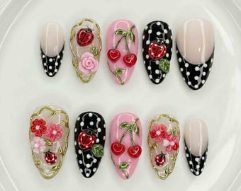 May include: A set of ten artificial nails with various designs. Some nails are nude with black polka dot tips. Others feature cherries, ladybugs, and flowers. The designs are set against a white background.