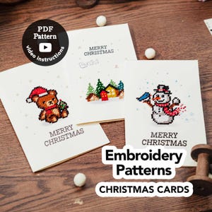 May include: Three white greeting cards featuring cross-stitch designs of a teddy bear in a Santa hat, a winter village scene, and a snowman. Each card displays the text MERRY CHRISTMAS. A black circular badge indicates PDF Pattern and video instructions.