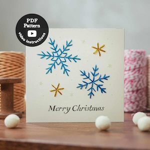 Könnte beinhalten: Eine quadratische, cremeweiße Grußkarte mit zwei gestickten blauen Schneeflocken und zwei kleinen goldenen sternförmigen Akzenten. Der Text Merry Christmas ist unten in Schwarz gedruckt. Ein schwarzes rundes Abzeichen zeigt PDF Pattern video instructions.