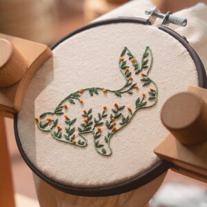 May include: An embroidery hoop with a cream-colored fabric featuring a green and orange floral bunny design. The bunny is outlined in green thread and filled with small orange flower-like details. The hoop is held in a wooden stand.