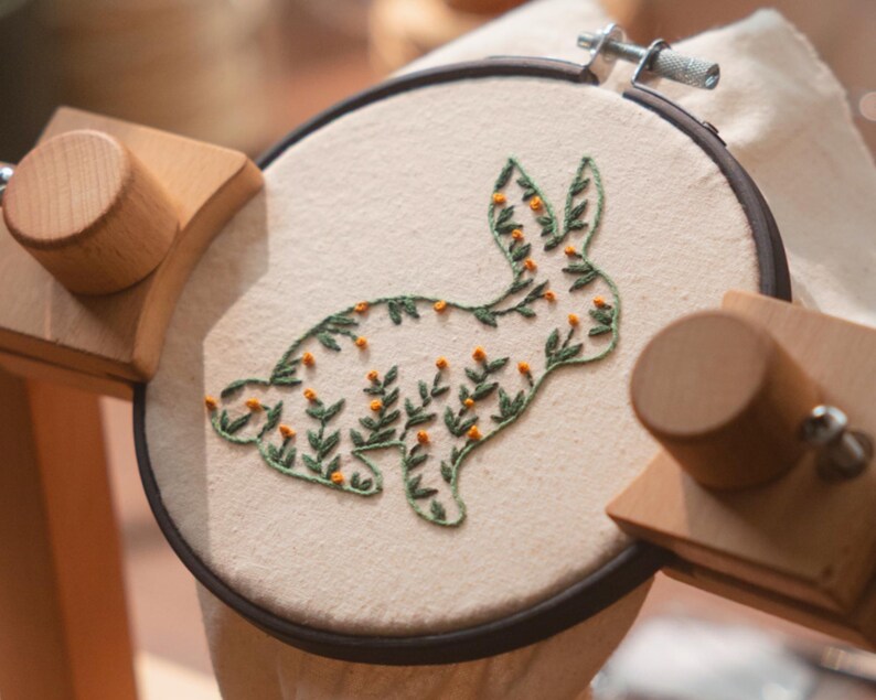 May include: An embroidery hoop with a cream-colored fabric featuring a green and orange floral bunny design. The bunny is outlined in green thread and filled with small orange flower-like details. The hoop is held in a wooden stand.