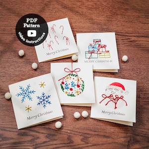Op de afbeelding: Vijf vierkante witte wenskaarten met geborduurde kerstontwerpen zoals zuurstokken, gestapelde cadeaus, blauwe sneeuwvlokken, een versierde kerstbal en een kerstman, elk met de gedrukte tekst Merry Christmas.