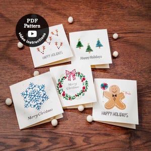 May include: Five square greeting cards featuring embroidered holiday designs including reindeer, Christmas trees, a snowflake, a wreath, and a gingerbread man. Each card displays the text Happy Holidays or Merry Christmas in black ink.