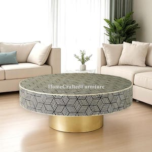 Può includere: Un tavolino rotondo con un motivo geometrico bianco e nero sulla parte superiore e sui lati. La base è dorata. Il tavolo è in un soggiorno con divani beige e una pianta.