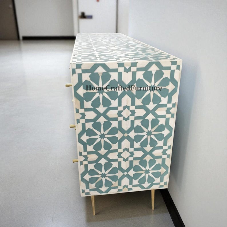 Puede incluir: Una c&oacute;moda con estampado geom&eacute;trico blanco y turquesa con tres cajones y tiradores dorados. La c&oacute;moda tiene patas doradas y el texto "HomeCraftedFurniture" es visible en el lateral.
