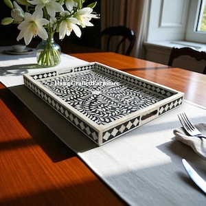 May include: Rectangular serving tray with a black and white floral design. The tray has a cream-colored border with diamond-shaped accents. The handles are cut out of the tray's frame. The tray is on a white runner on a wooden table.