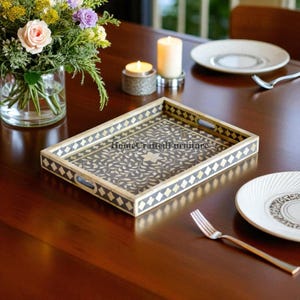 May include: A rectangular decorative tray with a gray and gold floral design. The tray has a diamond pattern border and handles. The text "HomeCraftedFurniture" is visible on the tray. The tray is on a dark wooden table, with flowers, candles, and tableware.