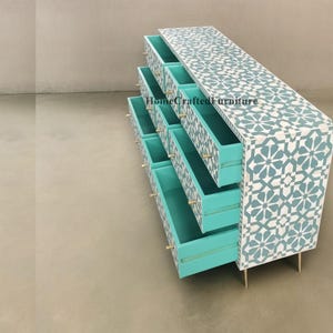 Puede incluir: Una c&oacute;moda baja y larga con m&uacute;ltiples cajones, con un patr&oacute;n geom&eacute;trico en blanco y azul claro. Los cajones est&aacute;n abiertos, revelando un interior turquesa. Las manijas y patas doradas completan el dise&ntilde;o. El texto "HomeCraftedFurniture" es visible.