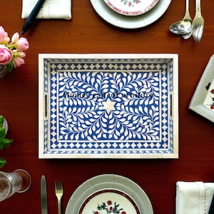 May include: Rectangular serving tray with a blue and white floral design. The tray has a white border and handles. The tray is on a dark wood surface, surrounded by tableware and flowers.
