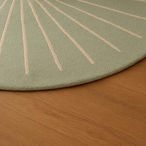 May include: A round, light green rug with a sunburst design in cream-colored lines. The rug is made of a textured material and sits on a wooden floor. The rug's design radiates from the center, creating a visually appealing pattern.