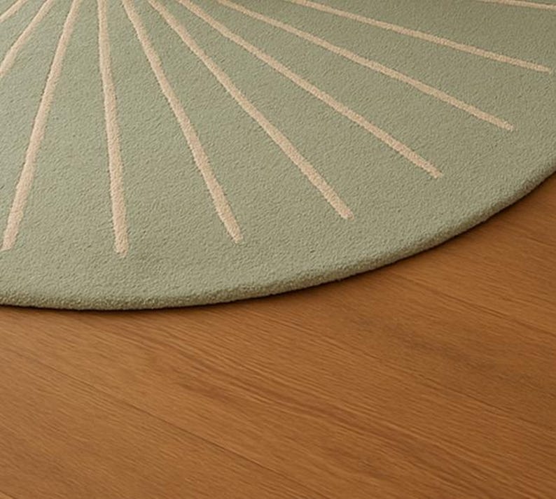 May include: A round, light green rug with a sunburst design in cream-colored lines. The rug is made of a textured material and sits on a wooden floor. The rug's design radiates from the center, creating a visually appealing pattern.